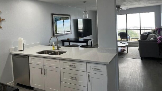 A modern kitchen with a central island, a dining area with a table and chairs, and a living area with a sofa and a view of the outdoors.