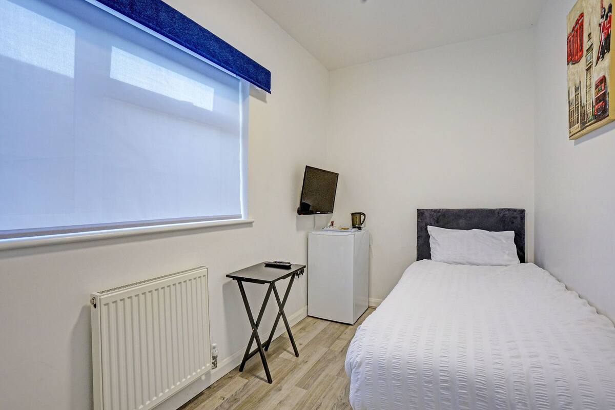 Ein Schlafzimmer mit Bett, Heizkörper, kleinem Tisch, Fernseher auf einem Schrank und einem Fenster mit Jalousien.