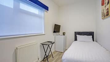 A bedroom with a bed, a radiator, a small table, a television on a stand, and a window with blinds.