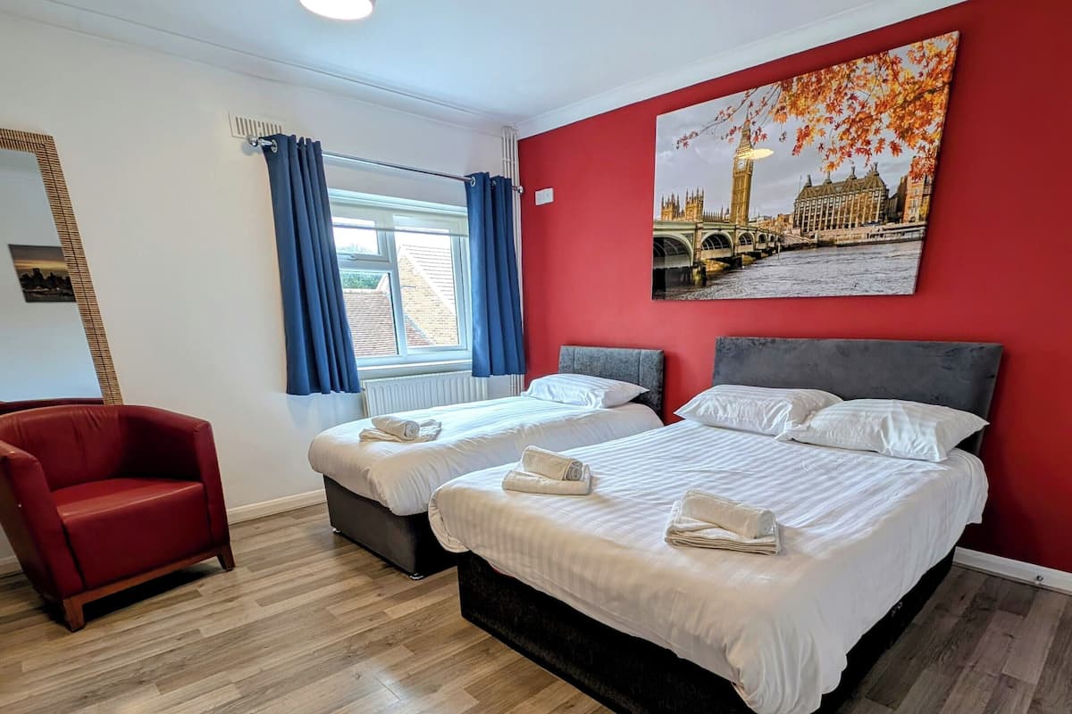 Ein Hotelzimmer mit zwei Betten, einem roten Sessel, einem Fenster mit blauen Vorhängen und einem Stadtbild an der Wand.