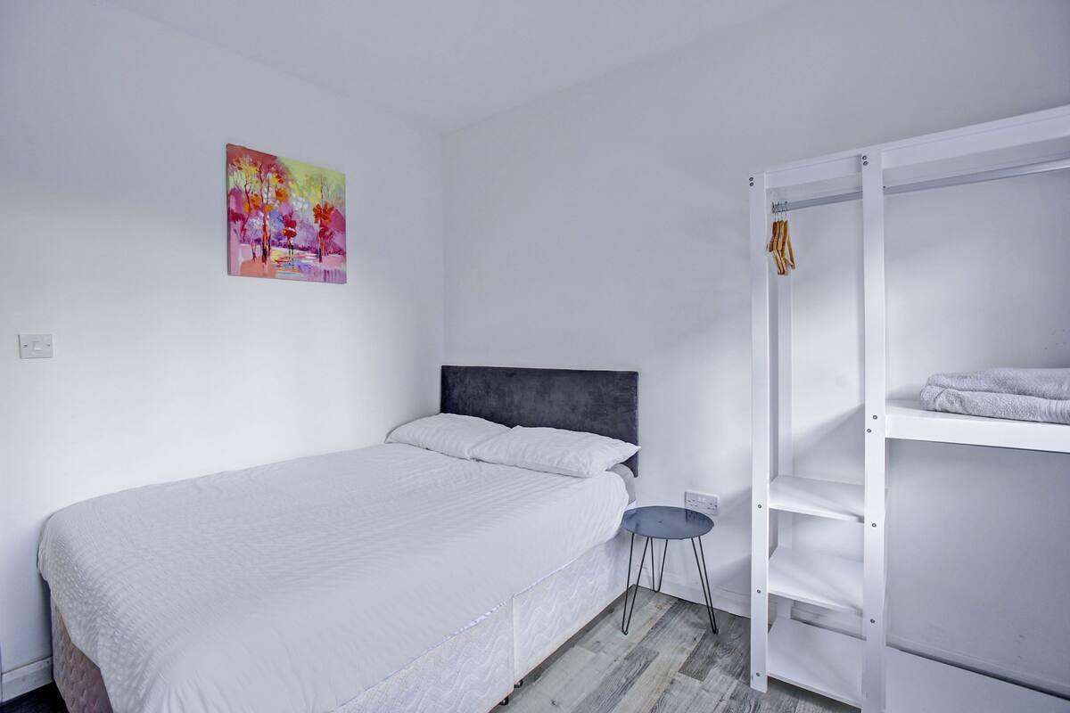 Ein Schlafzimmer mit Bett, kleinem Tisch, Stuhl, Kleiderschrank und einem farbenfrohen Bild an der Wand.