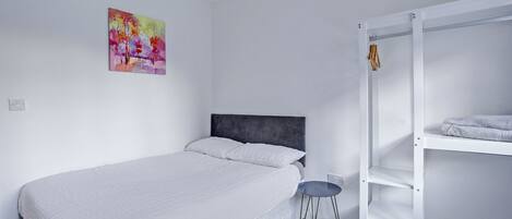 Un dormitorio con cama, una mesita, una silla, un armario y un cuadro de colores en la pared.