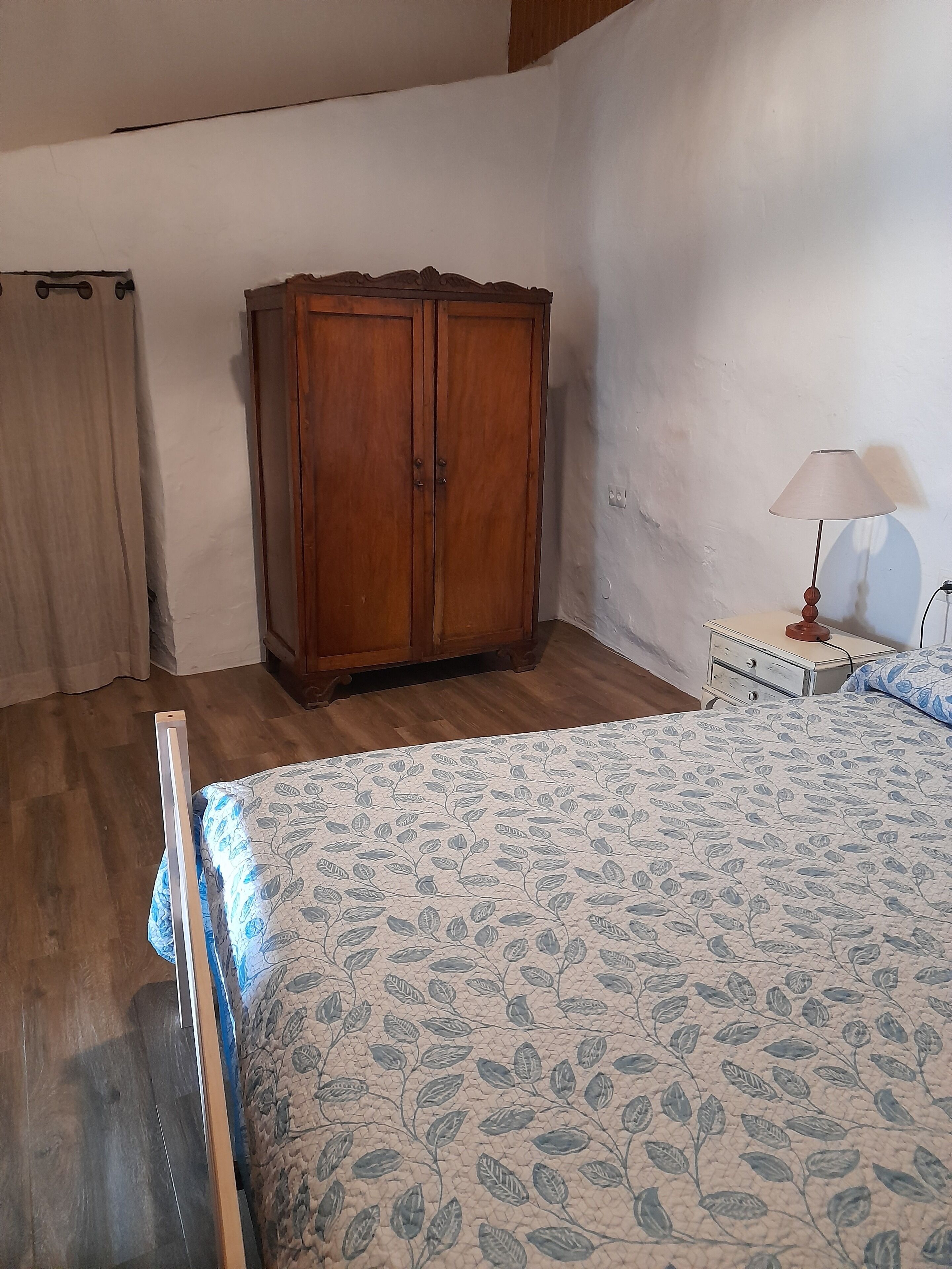 Un dormitorio con un armario de madera, una cama con colcha floral, una mesita de noche con lámpara y una ventana con cortinas.