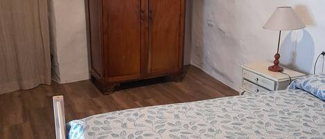 Un dormitorio con un armario de madera, una cama con colcha floral, una mesita de noche con lámpara y una ventana con cortinas.