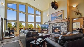 A spacious living room with a stone fireplace, leather sofas, and a large window overlooking a scenic view. - Native Retreat Cabin (Summit County)