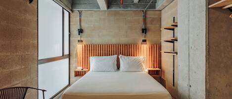 A narrow hotel room with a bed, a chair, and a view through a window.
