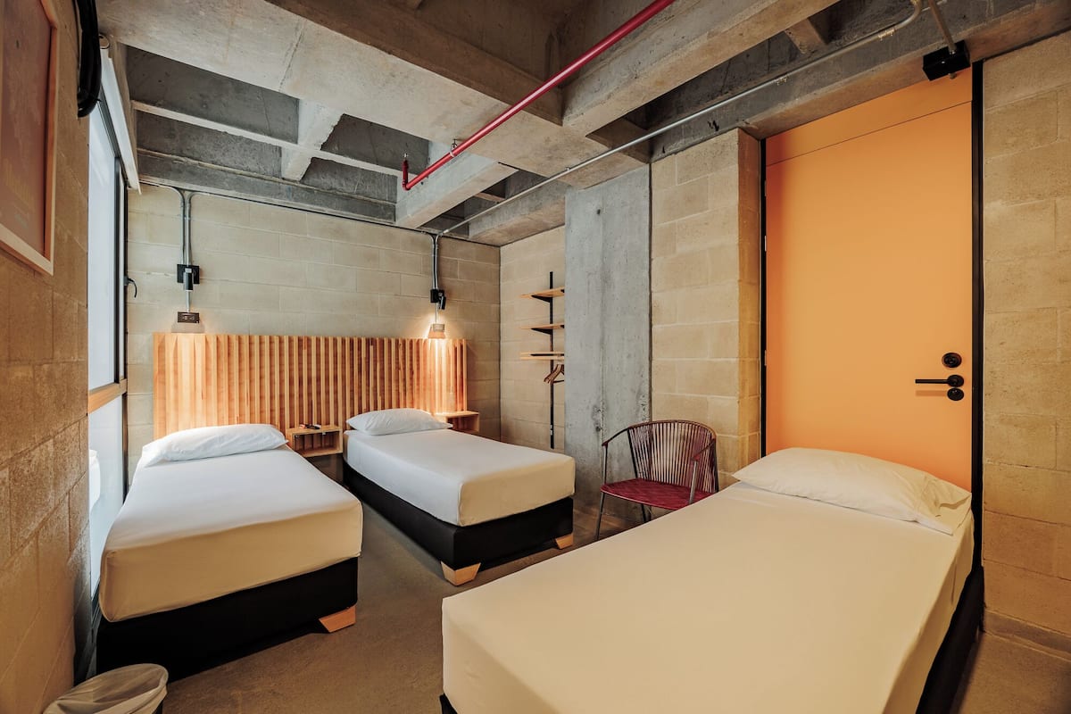 A room with two beds, a red chair, a wooden headboard, and a wall-mounted shelf.