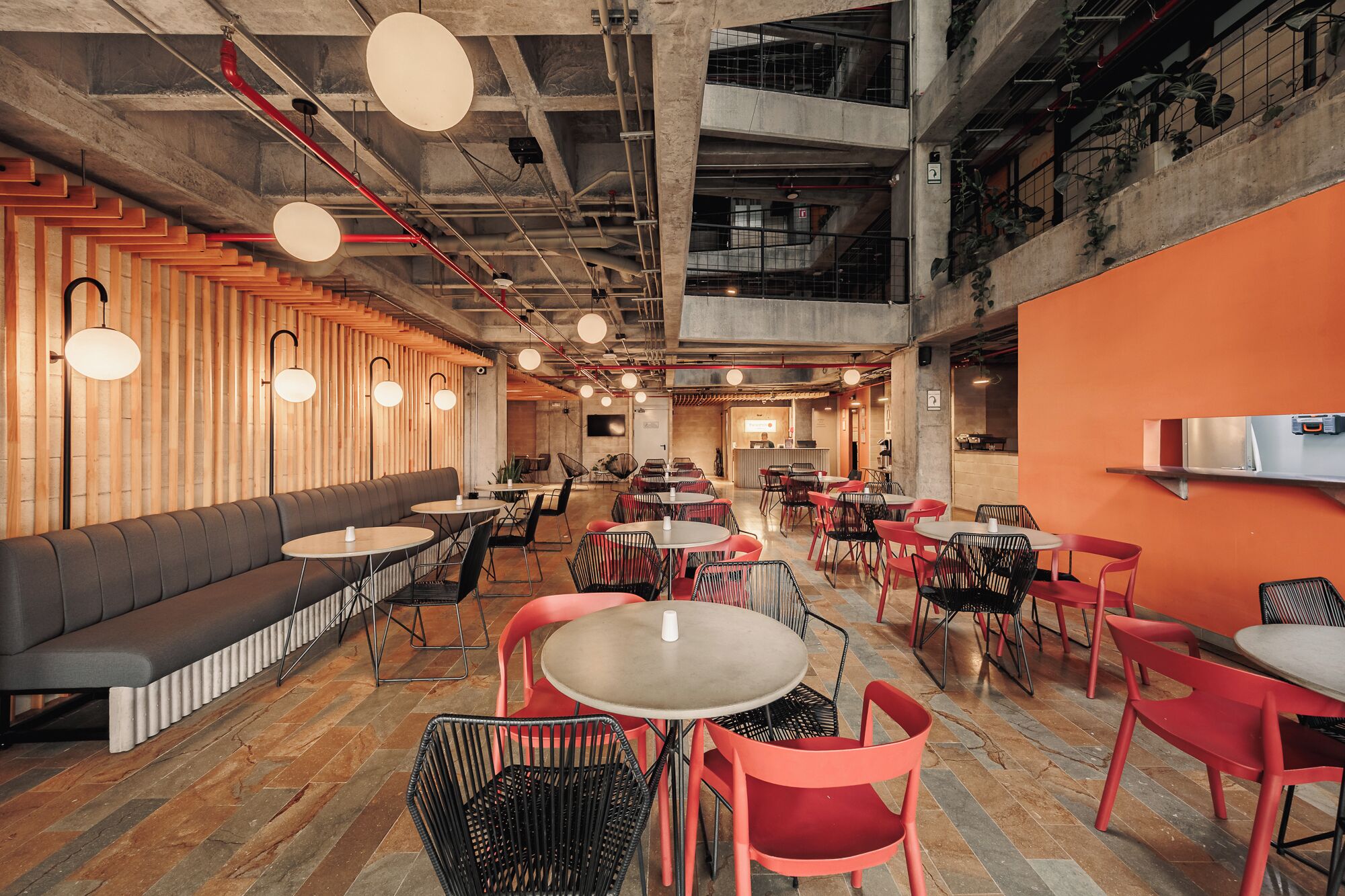 A modern interior with a mix of seating options including a long bench and round tables, a high ceiling with exposed pipes, and a combination of natural and artificial lighting.