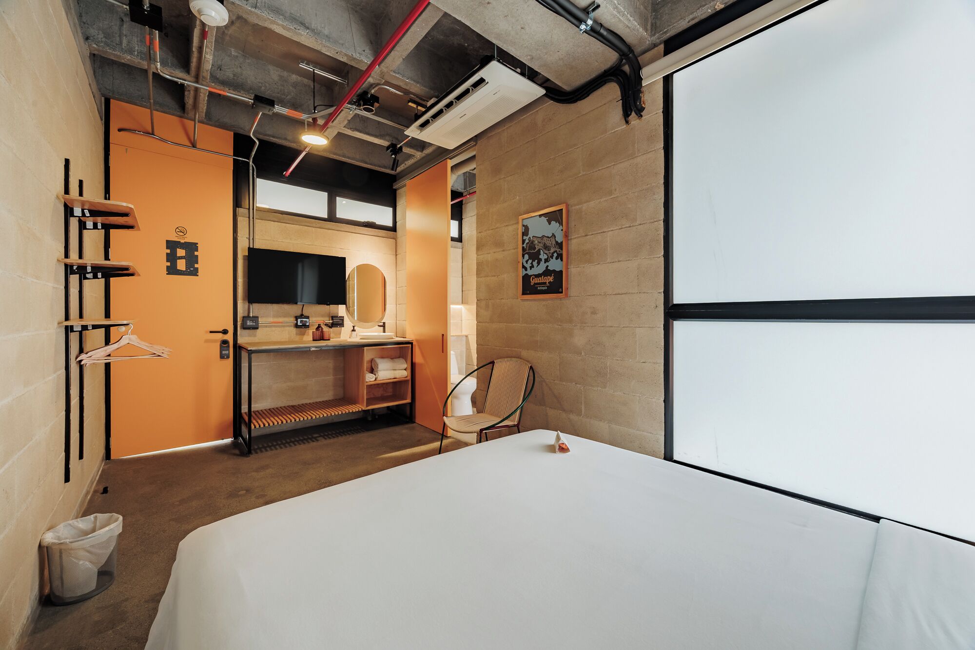 A hotel room with a bed, a desk with a mirror, a chair, a hanging coat rack, and a poster on the wall.