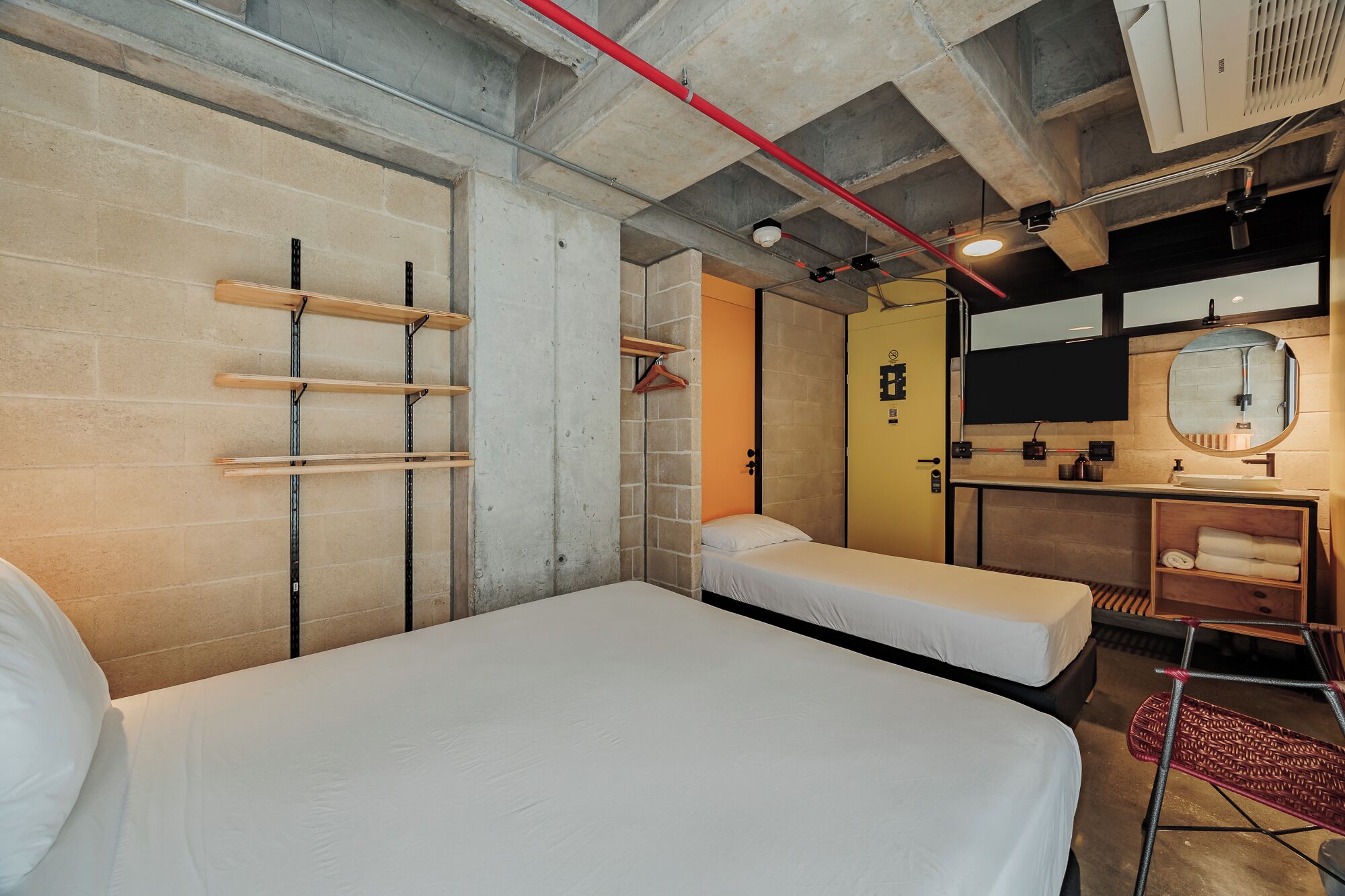 A compact hotel room with two beds, a bathroom with a mirror, and a concrete ceiling.