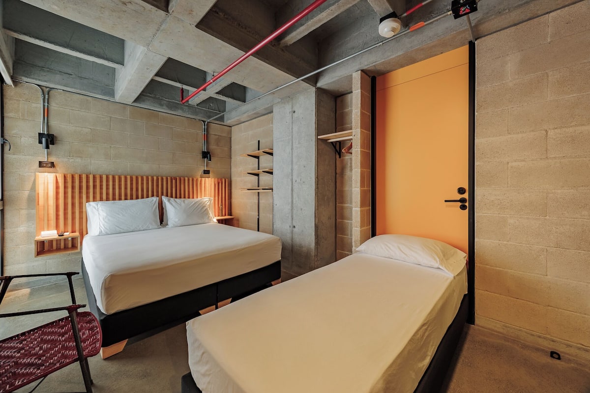 A modern hotel room with two beds, a concrete ceiling, and a brick wall.