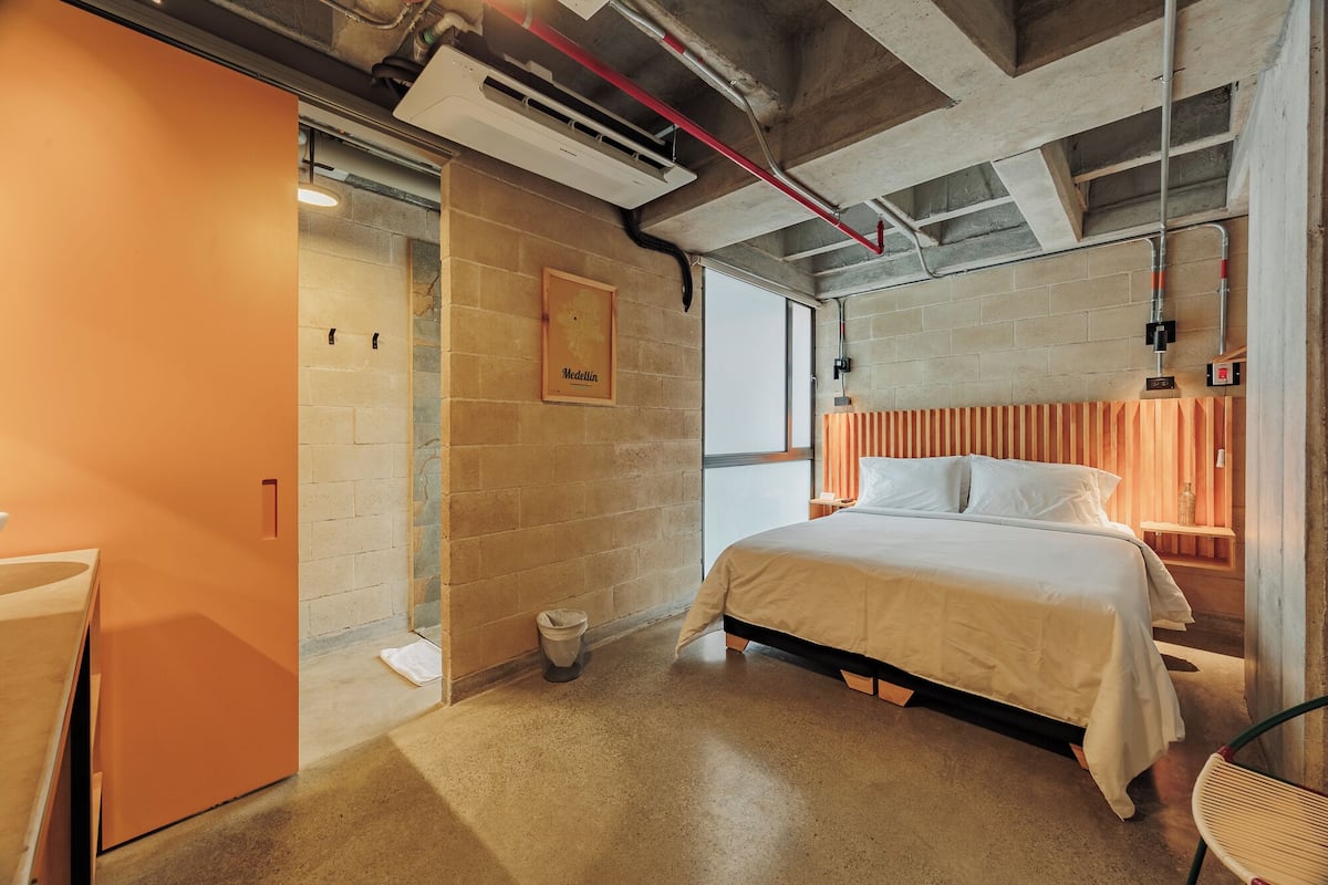 A bedroom with a bed, a wooden headboard, a wall-mounted air conditioning unit, and a visible window.