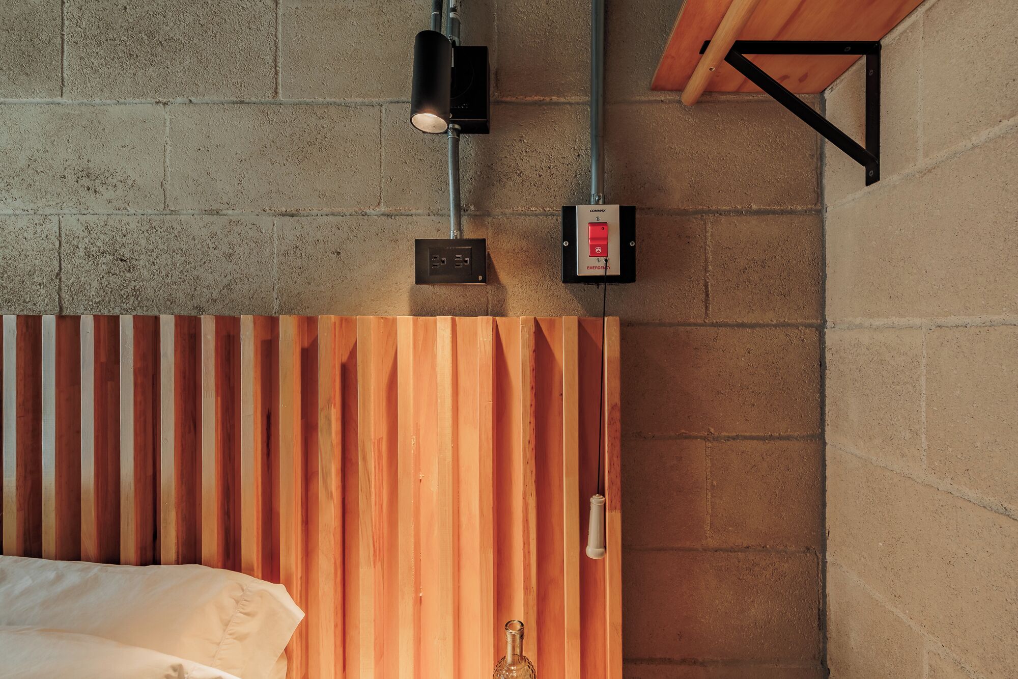 Une chambre avec une tête de lit en lattes de bois, un lit, une lampe de sécurité fixée au mur et une prise de courant.