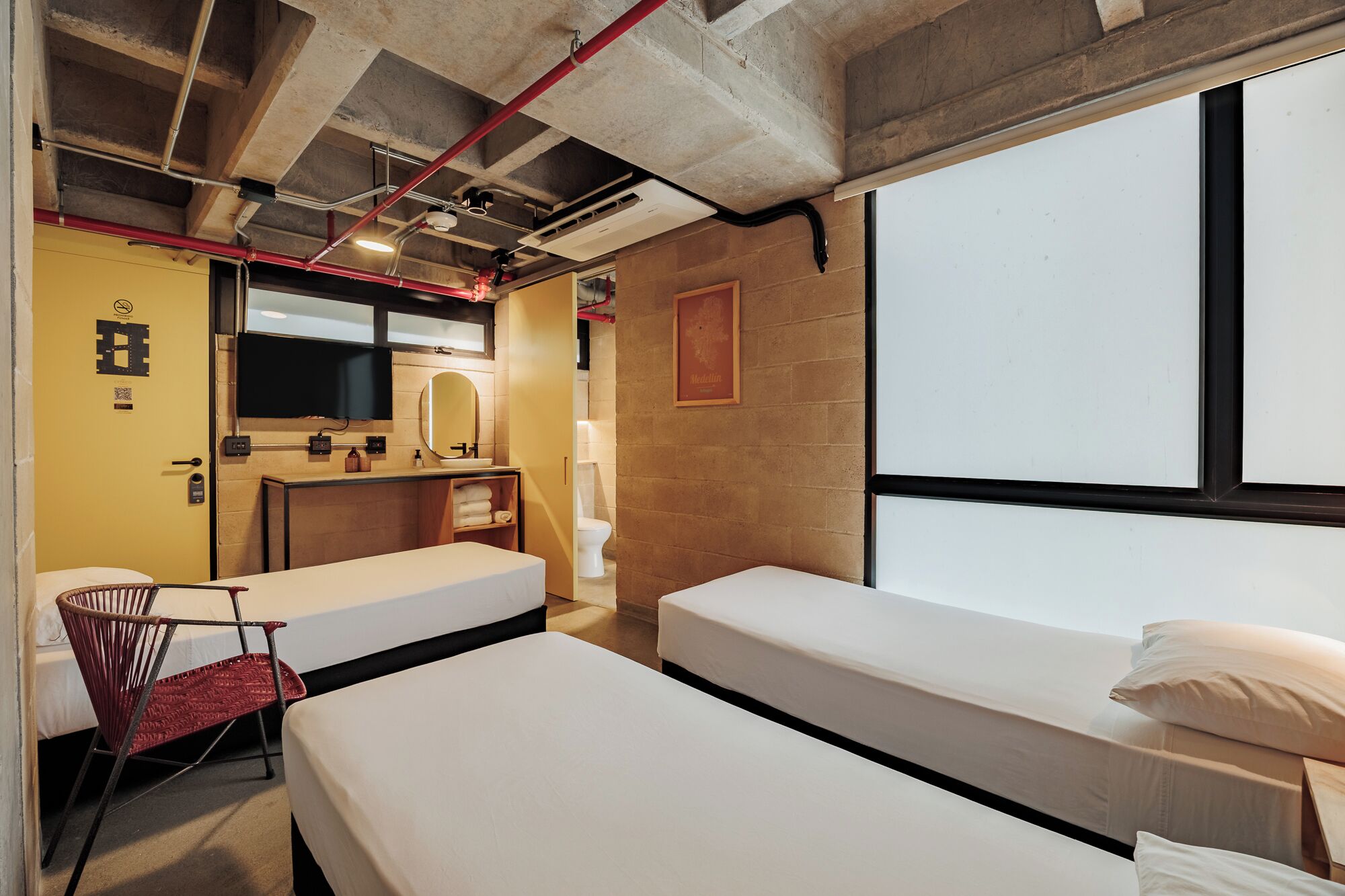 Ein Hotelzimmer mit zwei Einzelbetten, einem kleinen Schreibtisch mit Spiegel, einem Stuhl, einer Toilette und einem großen Fenster.