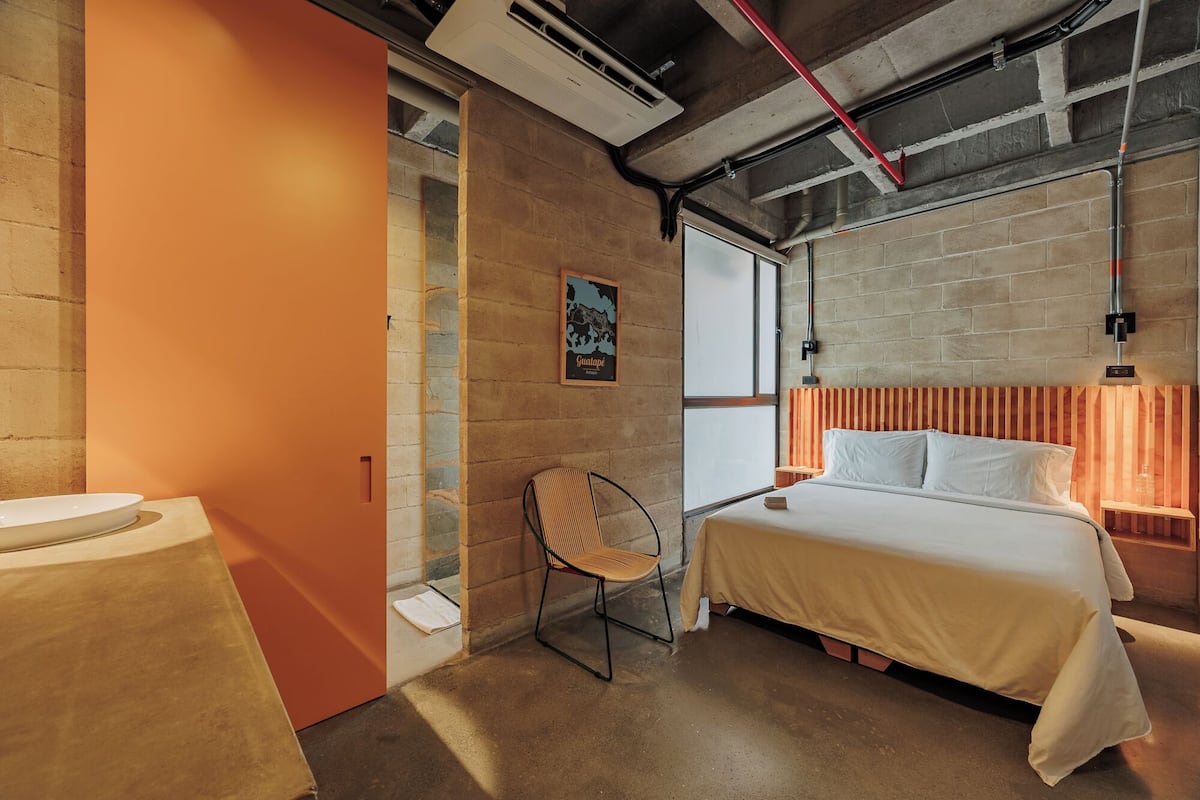 A hotel room with a bed, a chair, a toilet, and a window.