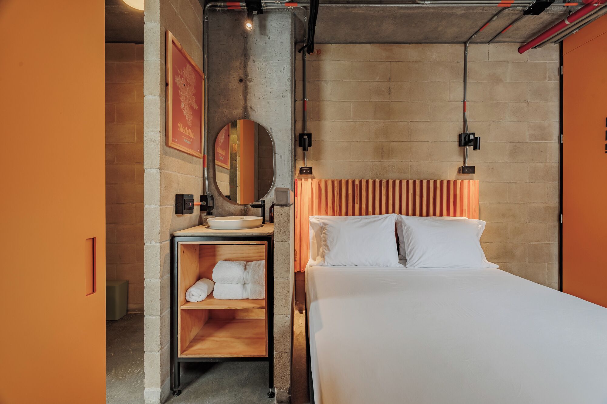 A bedroom with a bed, a wooden shelf with towels, a mirror, and a wall-mounted light fixture.