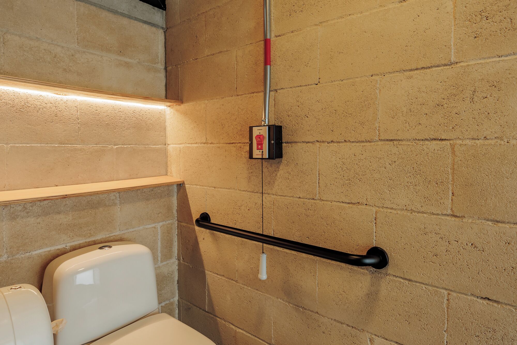 A restroom with a handrail, a wall-mounted emergency button, and a shelf with a toilet paper dispenser.
