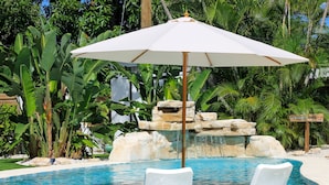 Un espace piscine agrémenté d’un parasol blanc, d’une cascade en pierre et d’une végétation tropicale luxuriante.