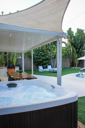 A hot tub under a canopy, surrounded by a wooden table and chairs, with a grassy yard and a wooden fence in the background. - A beautifully renovated private home with a large backyard, Pool, and Hot Tub (West Palm Beach)
