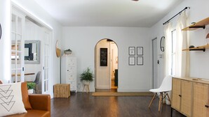 Un salon moderne avec un canapé en cuir marron, un fauteuil blanc et un meuble en bois. Une porte blanche donne accès à une autre pièce, et un petit tabouret en bois est placé à côté d’une plante.