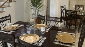 A dining area with a dark wooden table set for six, surrounded by black chairs with a geometric pattern. A staircase with wooden railings and a glass ceiling is visible in the background.