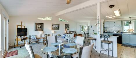 A spacious living area with a glass-top dining table, comfortable seating, a ceiling fan, and a kitchen area with light blue cabinets.