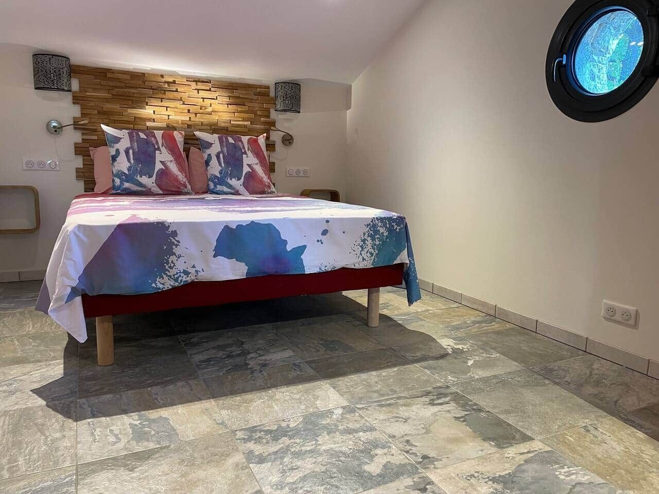 A bedroom with a bed, a wooden headboard, a round window, and a tiled floor.
