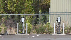 Zwei Ladestationen für Elektroautos auf einem Parkplatz, im Hintergrund ein Zaun aus Maschendraht und Bäume.