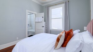 A bedroom with a bed, a window with curtains, a door leading to a bathroom, and a ceiling light fixture.