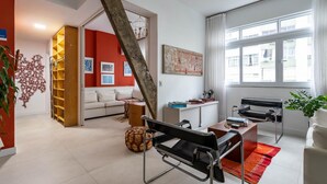 Un salon moderne comprenant un canapé, une table basse et un coin repas avec une table et des chaises. Une grande fenêtre offre une belle vue sur les bâtiments environnants et permet à la lumière naturelle d’entrer.