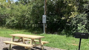Une table de pique-nique avec des bancs, entourée de gravier et d’herbe, avec une boîte aux lettres et un poteau électrique à proximité.