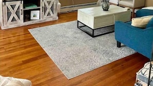 Un salon comprenant un téléviseur, une table basse, un fauteuil bleu, un escalier et une fenêtre avec des stores.