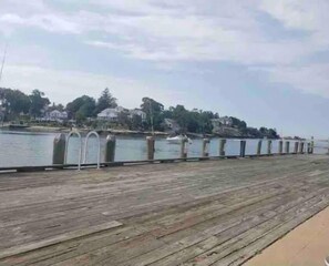 Un muelle de madera junto a una masa de agua, con casas al fondo.