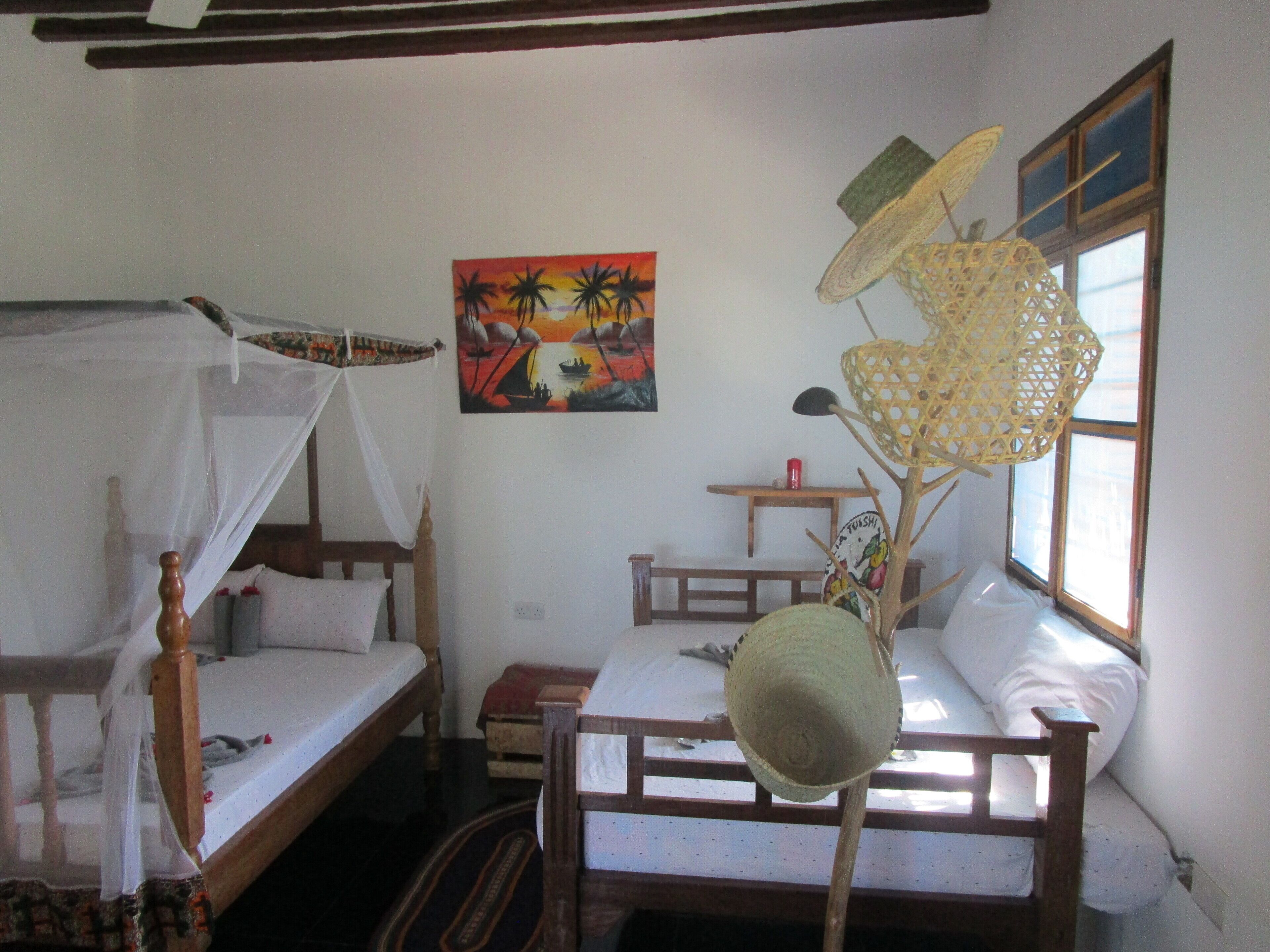 Una habitació amb dos llits de fusta, un amb mosquitera, un quadre d'un paisatge al capvespre i una cistella teixida penjant del sostre.