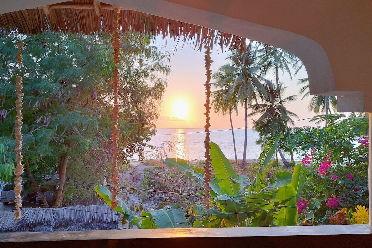 A view through a window with a thatched roof, tropical vegetation, and a sunset over the ocean.