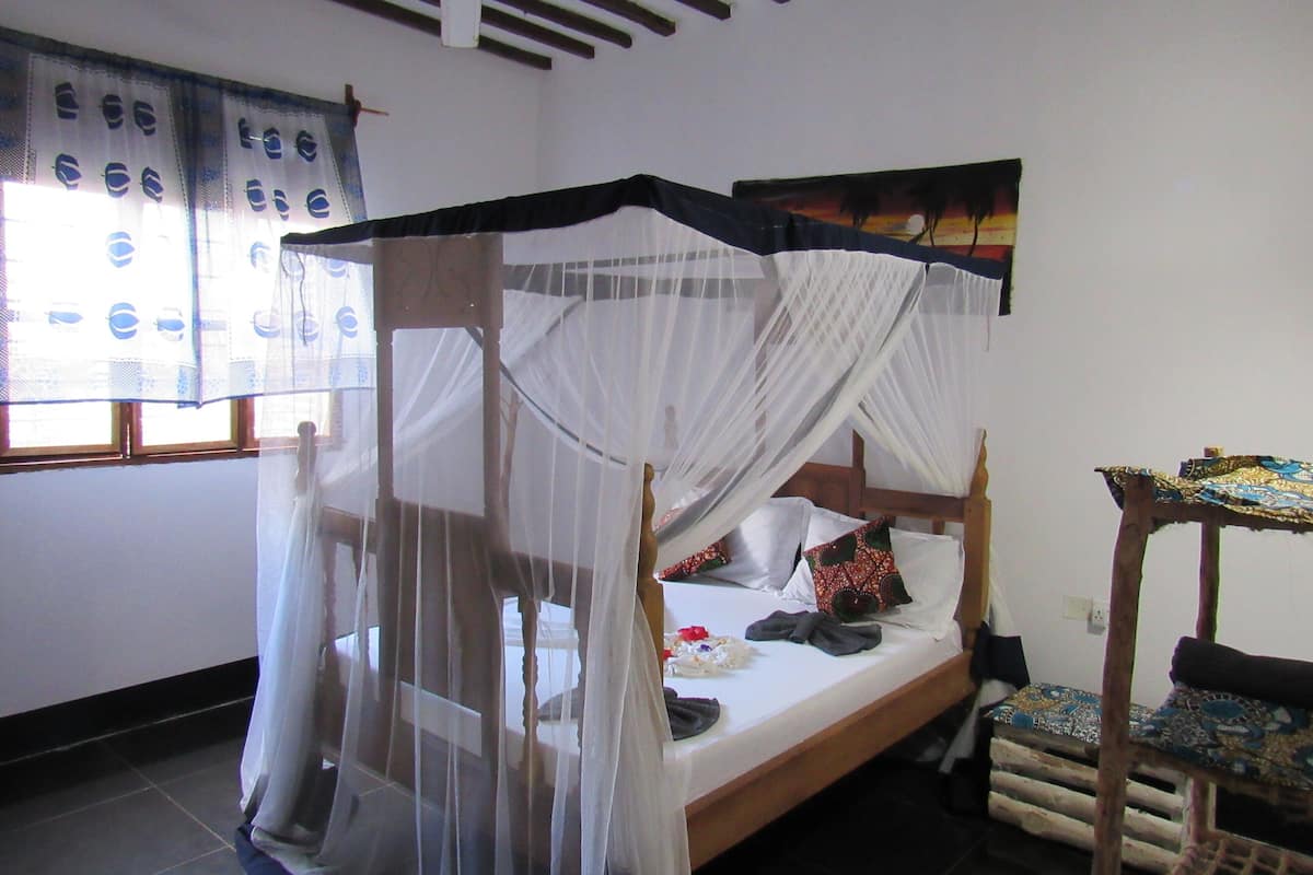 A room with a bed under a canopy, a small table with a patterned tablecloth, and a window with curtains.