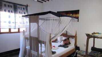 A room with a bed under a canopy, a small table with a patterned tablecloth, and a window with curtains.
