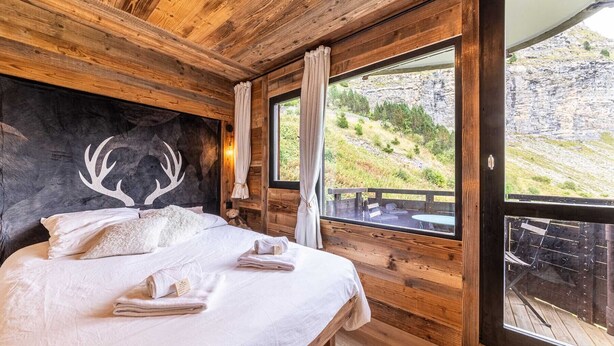 Une chambre à coucher dans un chalet en bois, avec un lit, une décoration murale représentant une tête de cerf et un balcon offrant une vue sur la montagne.