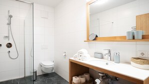 Une salle de bain moderne avec une douche à parois de verre, des toilettes, un lavabo avec un miroir au-dessus, et des étagères contenant des serviettes et des articles de toilette.