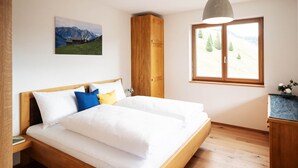 A bedroom with a wooden bed, a window with a view of trees, and a painting of mountains. - Landhaus Damüls - mountain view #08 (Damüls)