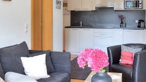 A modern living room with a grey sofa, a black leather chair, and a wooden coffee table with a vase of pink flowers. A kitchen with white cabinets and a microwave is visible in the background.