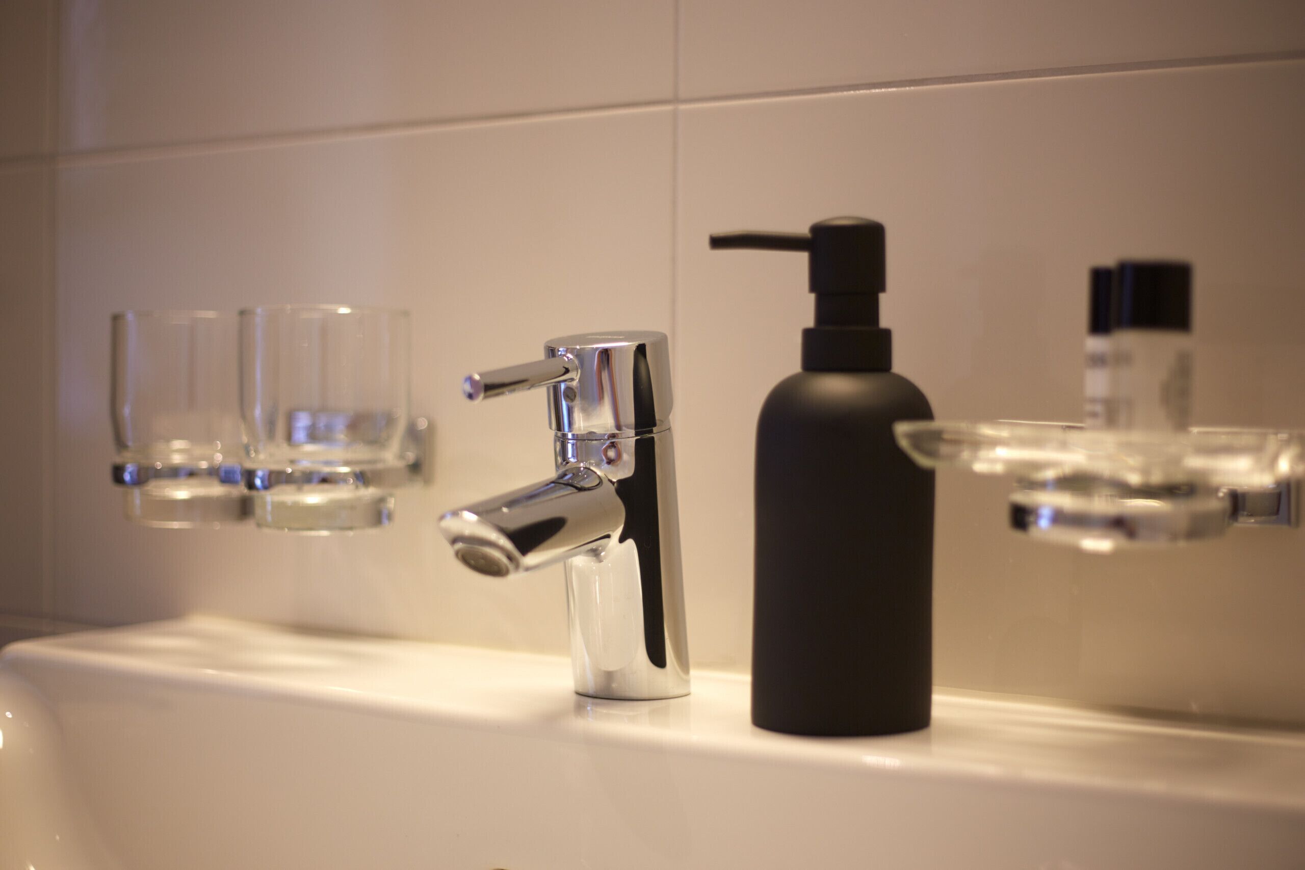 Een badkamer met een moderne kraan, een zwarte zeepdispenser en drie heldere glazen op de wastafel.