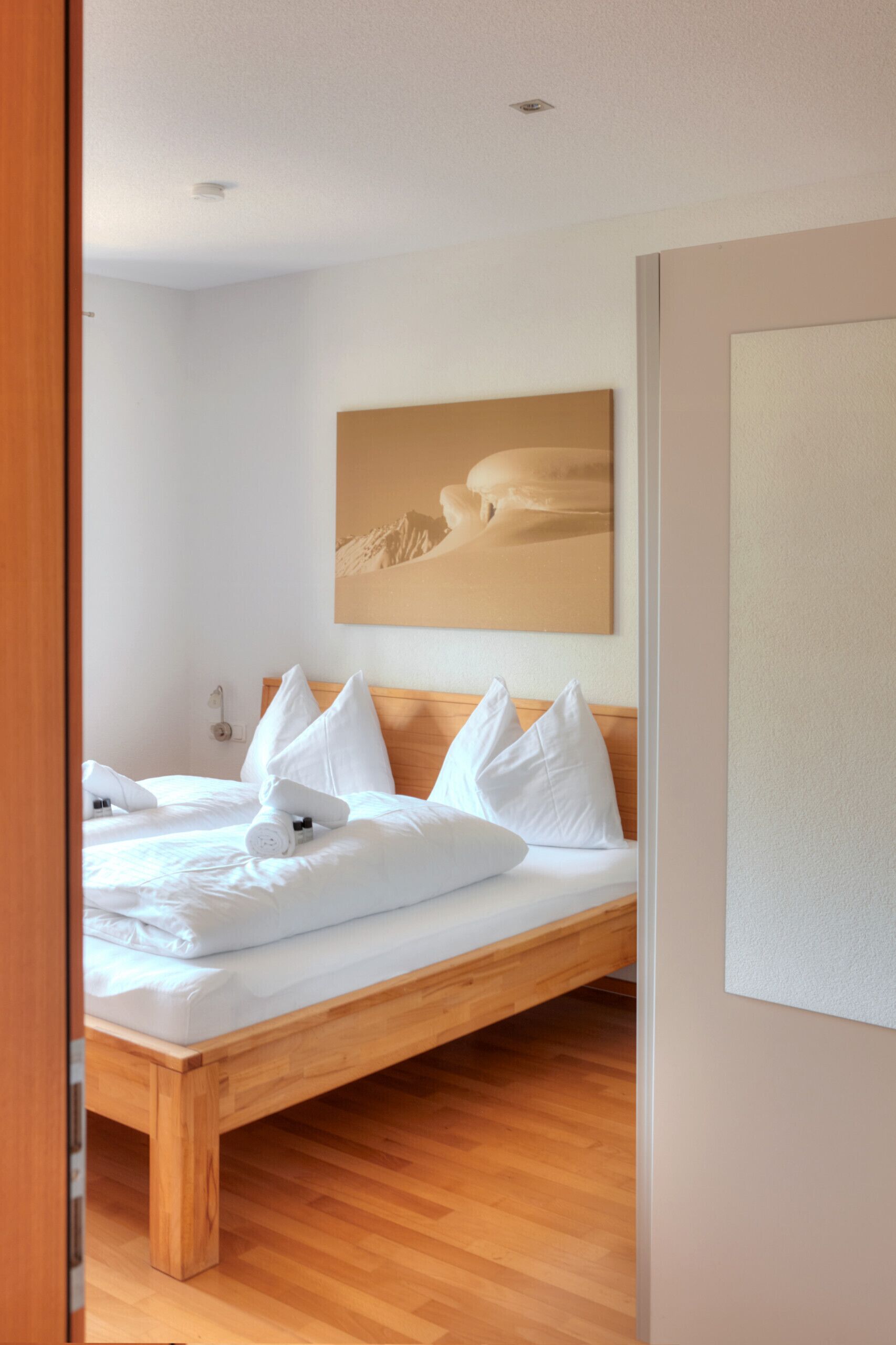 A neatly made bed with white linens and pillows, a wooden headboard, and a painting of a desert landscape on the wall.