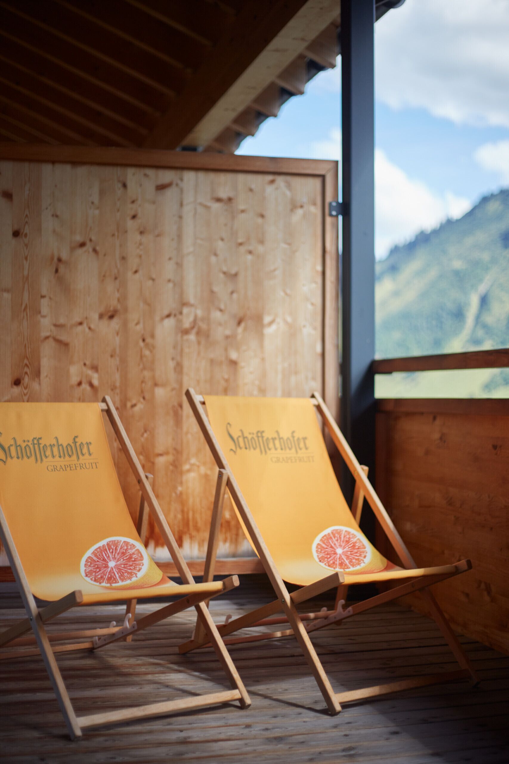 Two wooden lounge chairs with 'Schöfferhofer Grapefruit' branding, positioned on a wooden deck with a wooden wall and mountain view in the background.