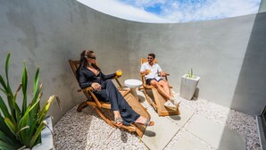 Dos personas sentadas en sillones de madera al aire libre, con un muro de concreto y una pequeña mesa entre ellas.