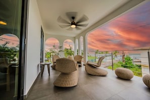 Uma área de estar moderna com móveis de vime, um ventilador de teto e vista para o pôr do sol através de amplas janelas.