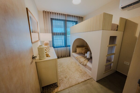 Quarto compacto com uma cama beliche, uma mesa de cabeceira com abajur e uma janela com cortinas.
