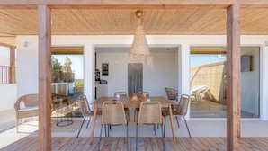 Un terrazzo in legno con tavolo e sedie, un lampadario e vista sull'interno attraverso porte in vetro.