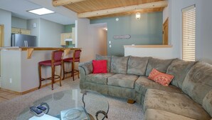 A living room with a wooden ceiling, a glass coffee table, a sofa with two cushions, and a kitchen area in the background. - Amalia Mountain Getaway w/ Deck, Fireplace! (Amalia)