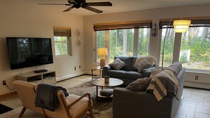 A living room with a grey sofa, a wooden chair, a coffee table, a TV, and large windows with a view of trees. - 4 Seasons Superior Beach House (Marquette)
